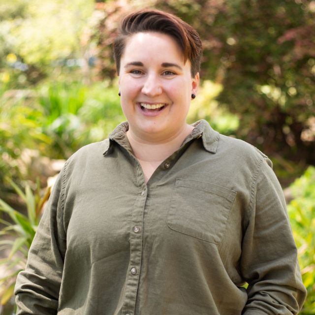 Headshot of Chelsea standing in a park. They have short brown hair and are wearing a green shirt and black jeans