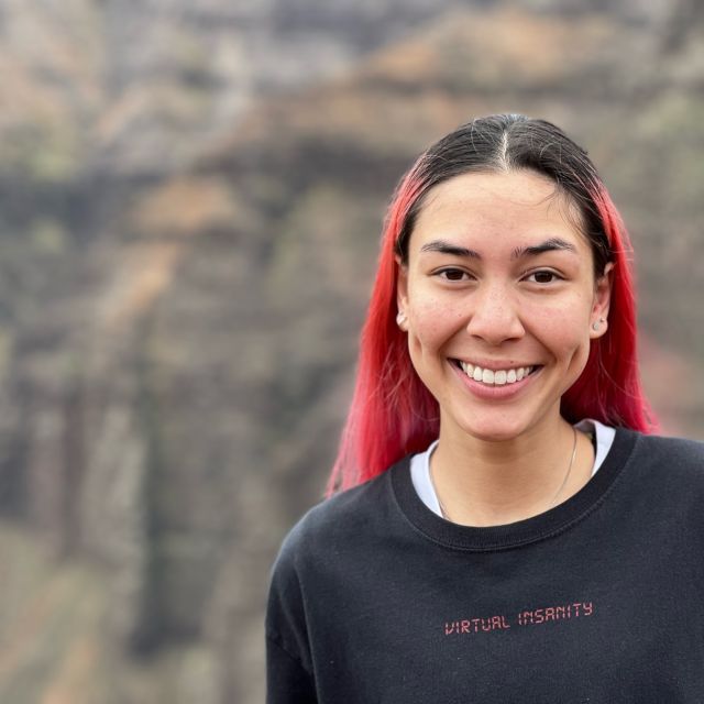 Headshot of Vanessa, she is standing outside, rocky mountain wall behind her, wearing a black shirt and has bright red dyed long hair.