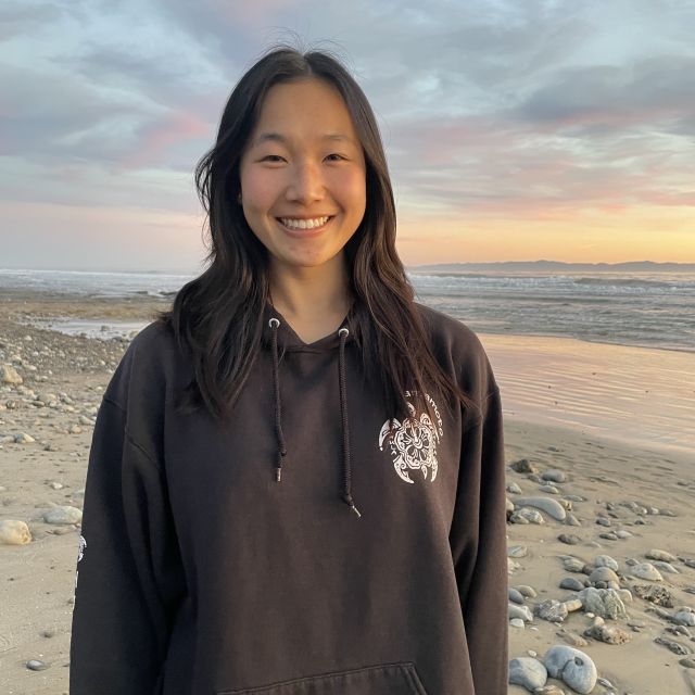 Photo of Ashley Fong wearing a black sweater standing at the beach during sunset